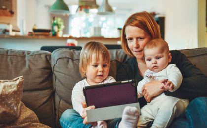 two babies and woman sitting on sofa while holding baby and watching on tablet