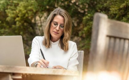 A woman in glasses writing and working on her laptop outdoors in natural daylight.